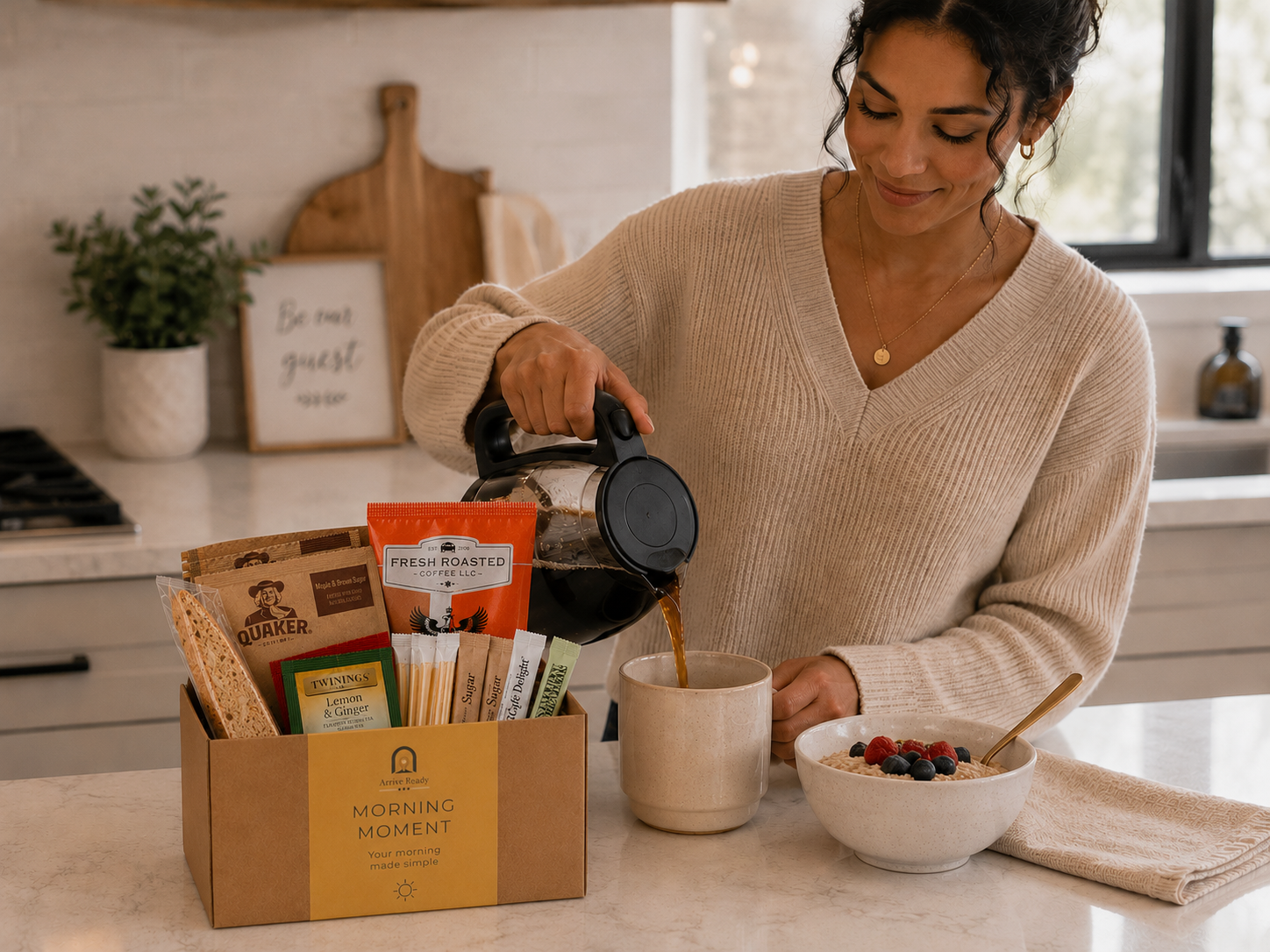 A woman in a beige ribbed sweater pours coffee from a glass carafe into a mug in a bright kitchen. In the foreground, a brown kraft gift box  Arrival kit labeled 'Arrive Ready MORNING MOMENT' contains assorted tea and coffee bags.