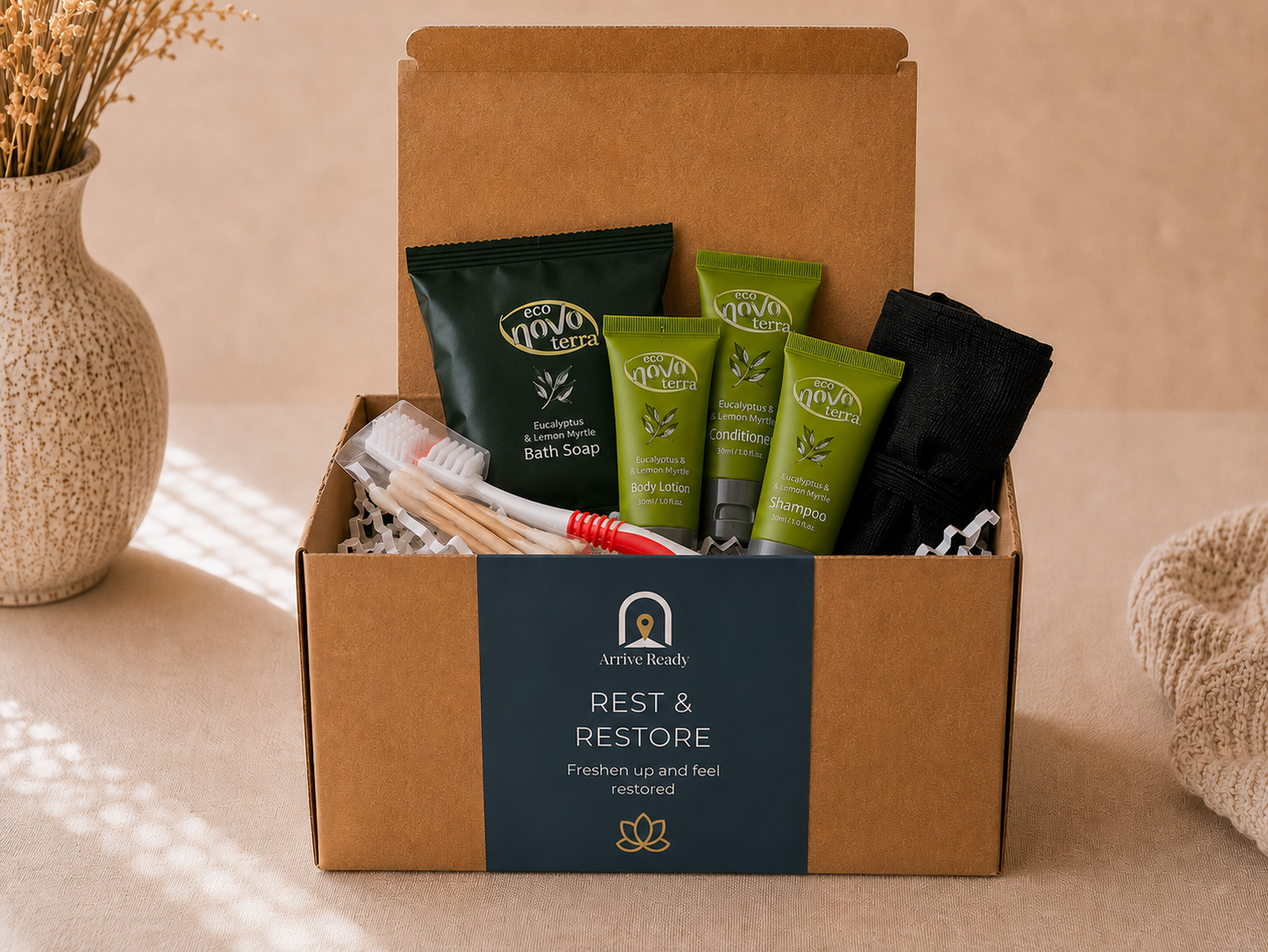 An arrival kit of a brown kraft gift box labeled 'ARRIVE READY REST & RESTORE' filled with bath and body items, including a dark green bath soap pouch, shampoo, conditioner, body lotion, body wash, toothbrush, toothpaste, set on a beige surface with a vase and towel in the background.
Freshen up and feel restored. 