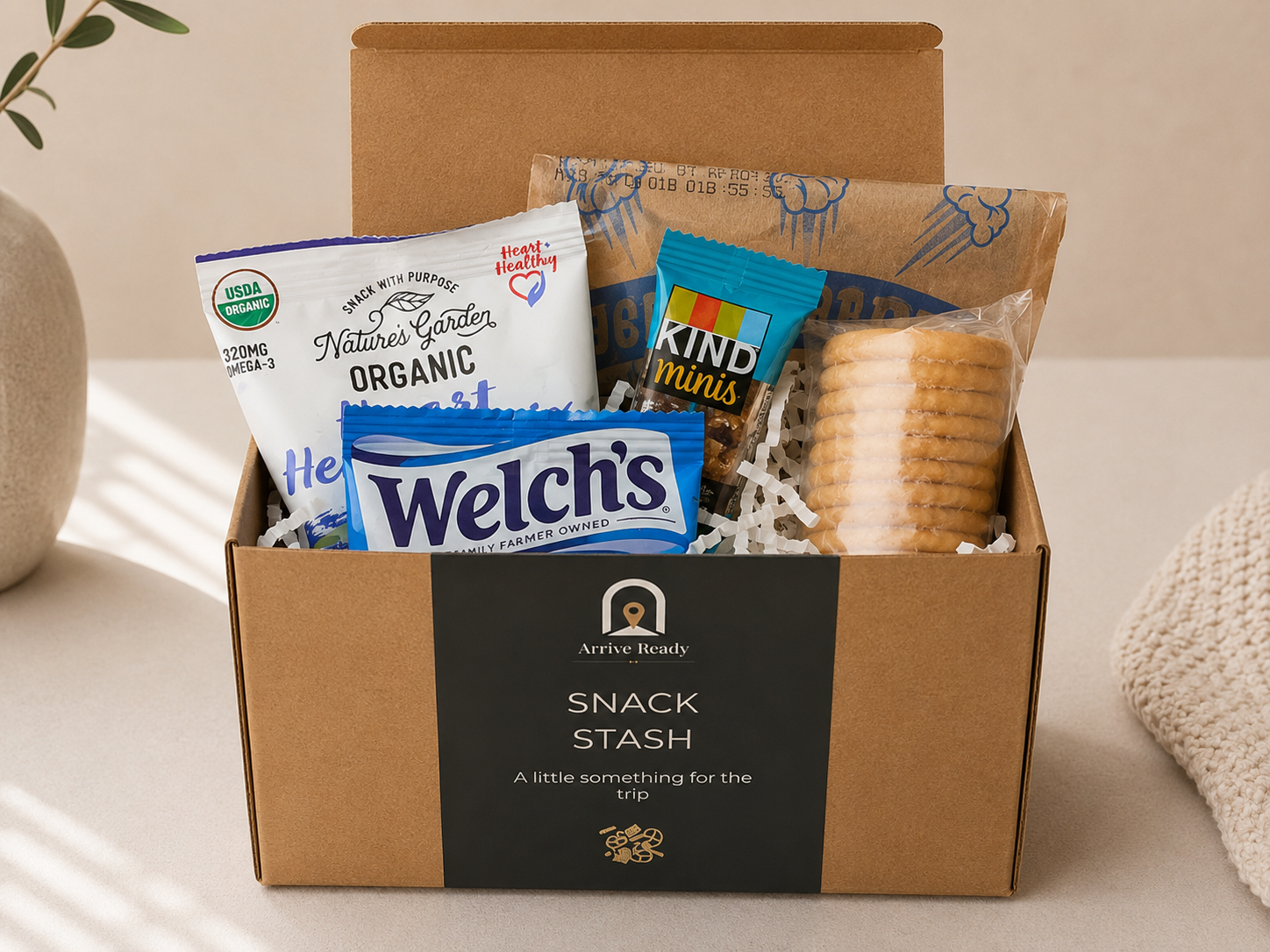 A brown kraft gift box arrival kit labeled 'Arrive Ready Snack Stash' filled with assorted snacks, including a Nature's Garden Organic snack bag, Welch's chocolate spread, and a blue KIND minis pack, set on a light beige surface. A little something for the trip. vacation rentals, travel, family