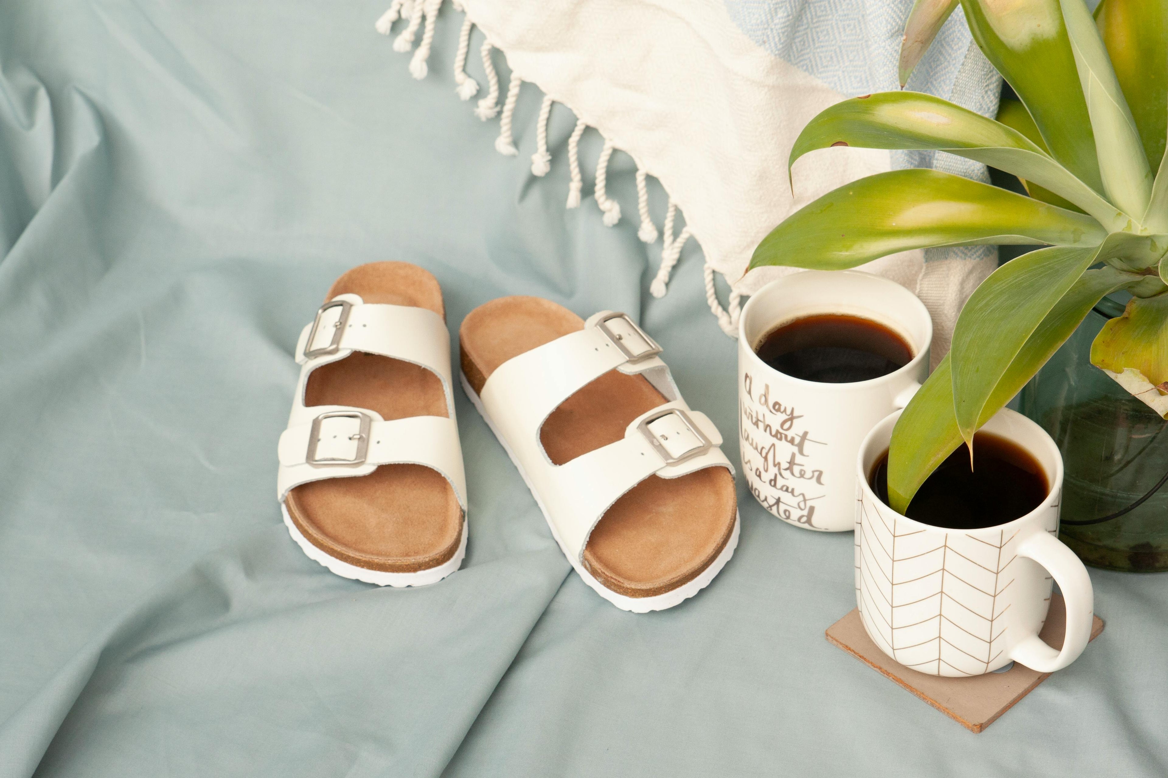 Pair of white sandals with brown soles on a light blue surface with two coffee mugs and a plant showing a relaxing arrival moment at vacation rental